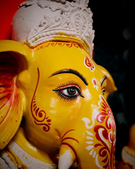 Close-up of a brightly painted yellow Ganesha statue typically associated with Hindu culture, featuring detailed facial features including large almond-shaped eyes with prominent black eyeliner and eyelashes, decorated with red and white ornamental patterns on the face. The statue's head is topped with an intricately carved white headdress or crown, and the image emphasizes the craftsmanship and vibrant colours. Although the image does not show a moving context or household items, it highlights cultural artifacts that could be involved in packing or transporting during a house relocation process. This detailed representation aligns with the theme of comprehensive packing and delivery services offered by [COMPANY_NAME], especially involving delicate or valuable items in a home moving situation. The focus on such culturally significant objects underlines the importance of careful handling during removals, ensuring safe transportation of all belongings, including decorative statues and art pieces, within a professional moving service environment as seen in the visual context.