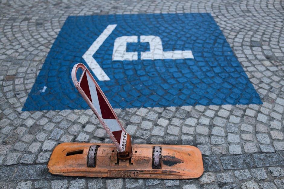 A close-up view of a cobblestone pavement featuring a painted blue and white accessible parking symbol indicating a designated parking space. In the foreground, there is a portable construction barrier with an orange base and a red and white striped warning sign leaning against it, suggesting temporary demarcation. The scene is captured outdoors in a public area, with natural lighting. This setting is relevant for house removals and home relocation logistics, illustrating parking regulations and temporary obstructions that may be encountered during furniture transport or loading and unloading processes. [COMPANY_NAME], a professional removals service, is familiar with managing such parking and access considerations during local moves, as shown in this environment supporting efficient packing, loading, or transportation activities.