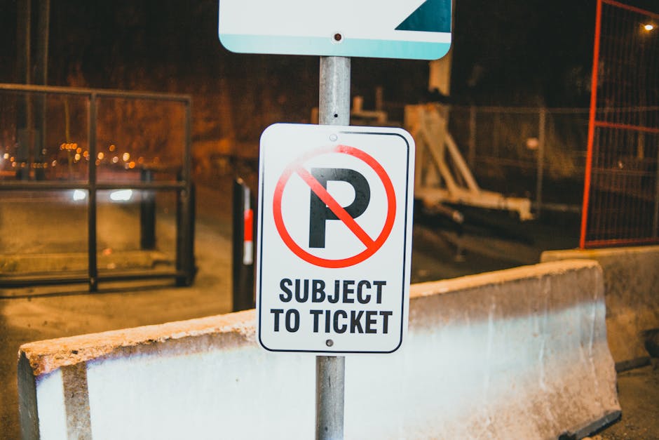 A metal signpost displaying a white rectangular parking restriction sign with a red circle and slash over a black 'P' symbol, indicating no parking, and the text 'SUBJECT TO TICKET' below. The sign is mounted on a metal pole situated behind a concrete barrier on a dimly lit street or loading area. In the background, there are blurred fence panels, a trolley, and some construction or storage materials. This scene likely depicts a designated unloading zone where house removal or furniture transport operations managed by [COMPANY_NAME] can take place under parking restrictions, with lighting suggesting an evening setting. The surroundings are utilitarian, consistent with areas used during home relocation or moving services, supporting safe and organized loading processes for furniture and boxes.
