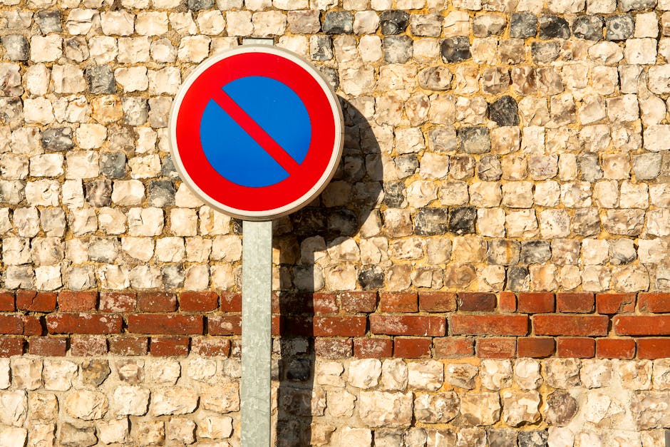 A close-up view of a cobblestone pavement featuring a painted blue and white accessible parking symbol indicating a designated parking space. In the foreground, there is a portable construction barrier with an orange base and a red and white striped warning sign leaning against it, suggesting temporary demarcation. The scene is captured outdoors in a public area, with natural lighting. This setting is relevant for house removals and home relocation logistics, illustrating parking regulations and temporary obstructions that may be encountered during furniture transport or loading and unloading processes. [COMPANY_NAME], a professional removals service, is familiar with managing such parking and access considerations during local moves, as shown in this environment supporting efficient packing, loading, or transportation activities.
