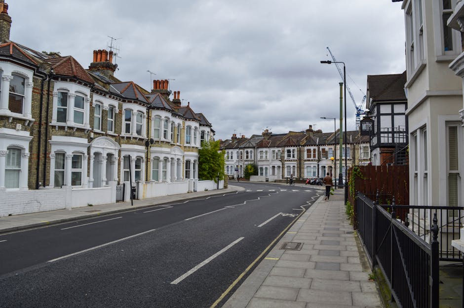 A quiet residential street in London featuring a row of Victorian-style terraced houses with brickwork facades, bay windows, and chimney stacks under a cloudy sky. The pavement runs alongside the houses, separated by black metal railings and small front gardens. There are parked cars along the curb and a single pedestrian walking down the sidewalk. The scene includes a streetlamp and a construction crane visible in the distance, suggesting ongoing development. This environment illustrates typical urban surroundings where house removals and furniture transport services by companies like [COMPANY_NAME] are often required for home relocation, involving loading and moving furniture, boxes, and appliances within similar streetscapes.