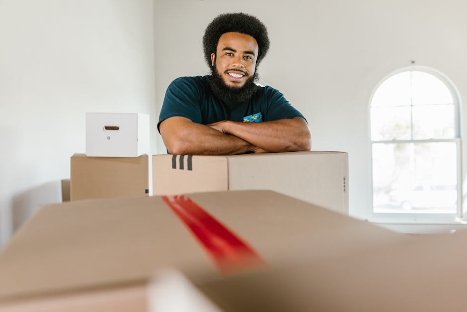 A smiling man with a dark complexion and curly hair, wearing a navy blue t-shirt, is standing inside a bright room with a large arched window in the background. He is leaning on a stack of moving boxes made of cardboard, which are arranged on a flat surface, likely within a home or apartment setting. The boxes vary in size, with some covered in packing tape, and one features a red stripe. The room has light-colored walls and natural light coming through the window. In the foreground, part of an open cardboard box is visible, indicating packing or unpacking activity. The scene reflects the process of home relocation, with packing and moving preparations underway, supported by the services of a professional removals company such as [COMPANY_NAME]. The environment appears organized for a house move, demonstrating proper handling of furniture and boxes, suitable for narrow access jobs like those on Walworth Road, as referenced in the page title, with equipment like trolleys or furniture blankets potentially used on the loading process.