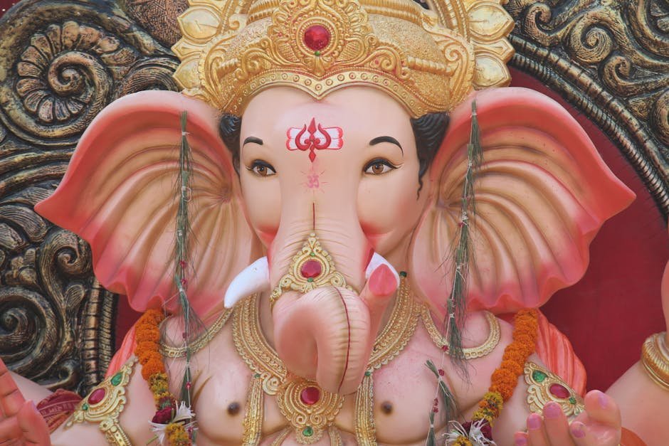 A detailed close-up of a large, colorful statue of the Hindu deity Ganesha, featuring an elephant head with pink ears, gold ornate jewelry, and a decorative crown topped with red jewels. The statue is adorned with various garlands and floral decorations, and is set against an intricately carved background with swirling patterns in dark tones. The lighting highlights the smooth texture of the statue’s face and the delicate craftsmanship of the jewelry and crown. This image is related to house removals and moving services provided by [COMPANY_NAME], illustrating the careful packing and handling of delicate or culturally significant objects during a home relocation through furniture transport or packing and moving processes.